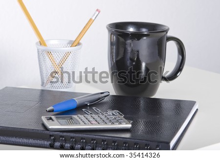 Calculator and pen sitting on a folder with pencils and a coffee cup in the background.