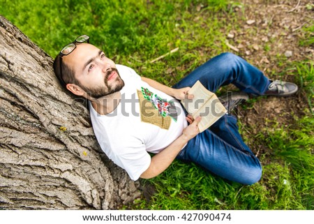 Young relaxed man reading book in nature, back on tree, meadow behind