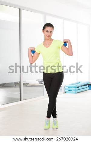 Beautiful woman with dumbbells posing for camera in sport gym. Woman weared in yellow t-shirt and black jogging trousers.