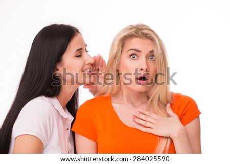 Young pretty girlfriend in orange T-shirt looking surprised. Friend of her in white T-shirt telling her something in a whisper.
