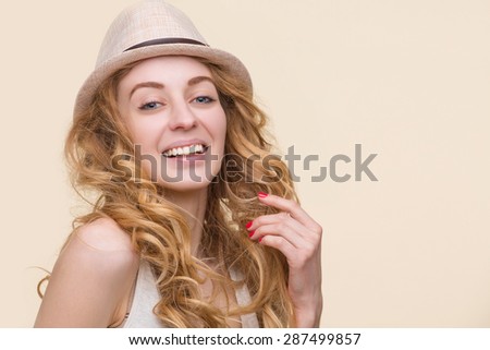Smiling young beautiful lady touching her red-coloured hair. Girl with red nails posing with cream hat on in photostudio.