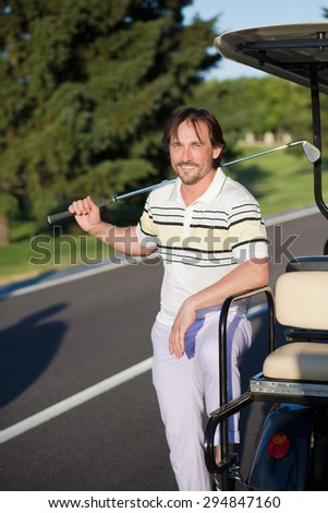 Mature golf player smilling near white gold car. Man in T-shirt and sports trousers keeping brassie in his right hand.