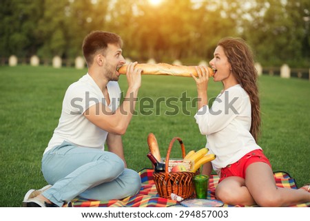 Young couple eating in picnic in beautiful sunset. Man and woman sitting face to face on carper, basket between them.