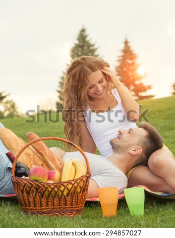 Young couple relaxing in picnic in the sunset. Man lying on his girl-friend\'s knees and smiling to her, basket and cocktails standing near.