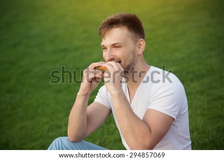Handsome man eating sandwich in picnic. Short-haired man in white T-shirt having a snack in beautiful sunset.