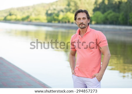 Rich man posing for photographer with his arms in pockets isolated on river. Middle-aged man in coral T-shirt owing huge factory.