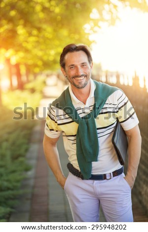 Handsome man standing with his hands in pockets. Smiling rich man posing with fashionable haircut in the park.