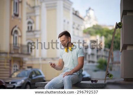 Man sitting in the centre of European city. Short-haired man in light T-shirt with yellow earphones searching telephone number.