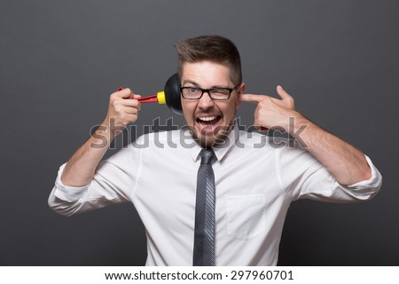 Exhausted man is angry after his work. Businessman in glasses keeping plunger in his ear and shouting so much isolated on grey.