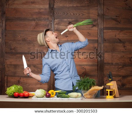 Friendly chef preparing vegetables in his kitchen. Handsome man in blue shirt going to cut leek with knife isolated on wooden.