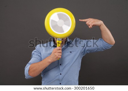 Chef hiding his face behind yellow pan and pointing out isolated on grey. Young man in blue shirt is professional cookery specialist.