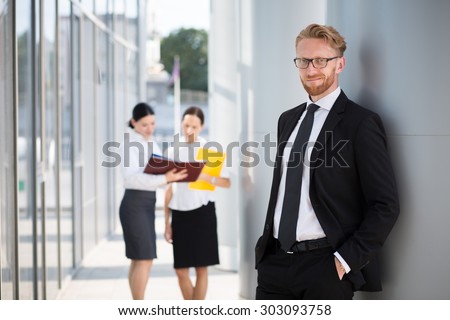 Happy smart businessman with team mates discussing on the background. Man in business suit smiling with his hands in pockets.