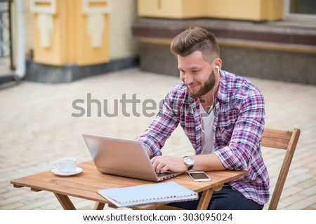 Photo of a thoughtful young man communicating with friends using Skype on laptop. Bearded man in earphones enjoyimg time in the cafe with cup of coffee.