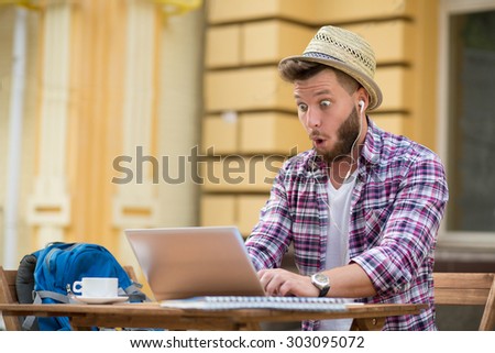Astonished and surprised man in straw hat playing computer games during lunch time. Man in earphones sitting in the cafe and drinking a cup of coffee.