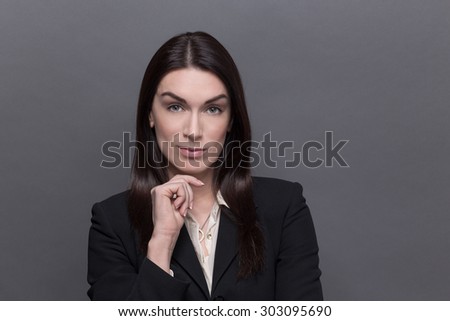 Portrait of business lady in black suit keeping her chin. Woman with long black hair looking so seriou and industrious isolated on grey.