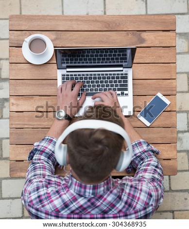 Close-up portrait of man freelancer working on laptop at wooden table. Man with earphones on typing smth and drinking hot coffee.