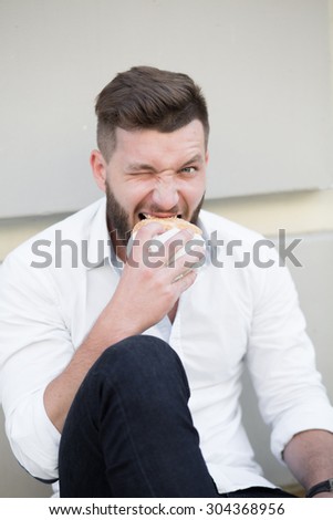 Handsome bearded man eating hamburger outdoors. He is hungry and having a good bite. Businessman resting during lunch time.