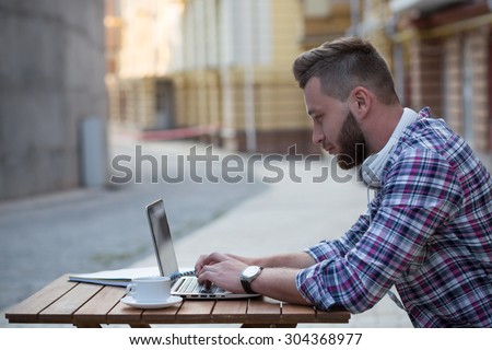 Man freelancer working over laptop in the evening. Serious hipster looking at the screen, typing the document and drinking coffee.