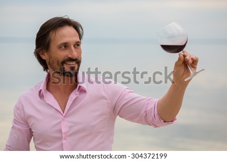 Close-up portrait of winemaker tasting red wine in glass. Handsome bearded man in pink shirt smiling and looking at wine in glass isolated on sea.