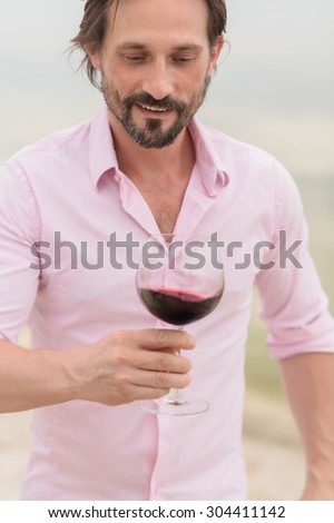 Confident and happy sommelier. Bearded mature man in pink shirt tasting red wine and looking at the glass isolated on nature.