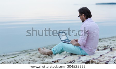Mature freelancer working on laptop. Author in glasses writing poem at the beach. He is becoming inspired by the beautiful sea.