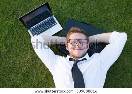 Tired freelancer lying in the park near his laptop computer. Handsome man in glasses resting with his hands on the nape.
