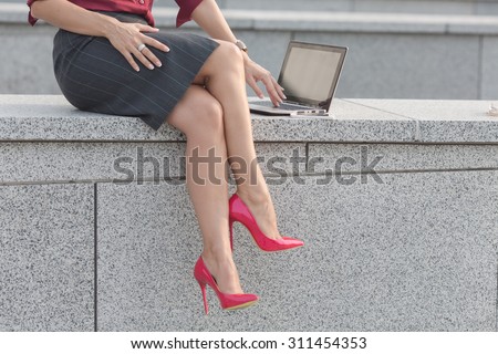 Close-up picture of beautiful woman\'s lags sitting on concrete brick. Lady in high heels working with laptop computer outdoors.