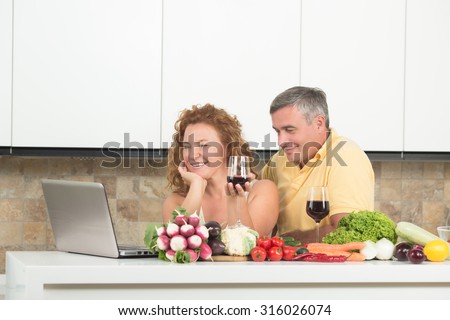 Happy mature couple watching videos on the laptop computer and drinking red wine. People cooking dinner or lunch in the kitchen.