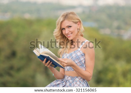 Happy smiling lady is in search of interesting moment in her best book. Beautiful woman dress reading book in summer park.