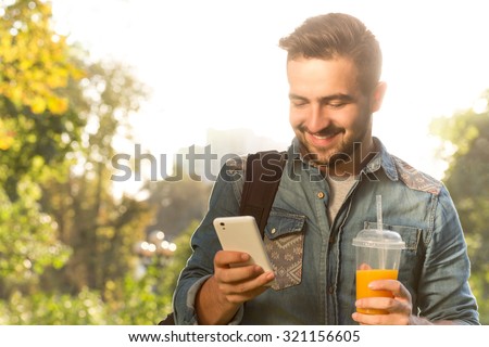 Happy hipster man using mobile phone and drinking orange cocktail. Handsome man spending his daytime walking in the park.