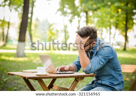 Freelancer hipster man working in the park at the table. Exhausted man searching something ib the Internet using his laptop computer.