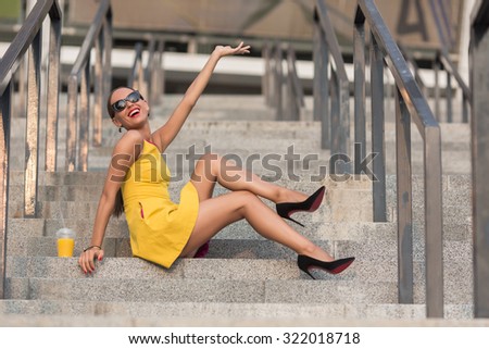 Toothy laughing fashion girl sitting on stairs of office building with orange cocktail. Lady with red lips raising hand to the sky.