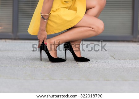Close-up portrait of woman\'s legs on high heels. Lady in yellow dress sitting and touching her right leg near office building.