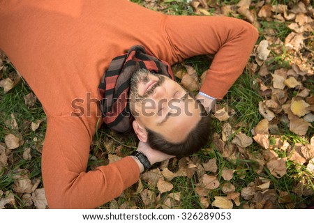 Handsome middle-aged man lying on the autumn leaves and resting from hard-working day in the park. Man in orange sweater lying with closed eyes.