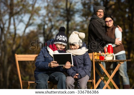 Family, autumn and picnic concepts. Happy little children sitting all together with tablet PC while their parents hugging on the background and looking at them.