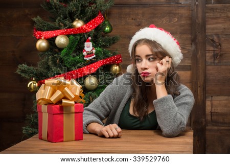 Sad girl sitting at the table and thinking about real holiday. Serious and excited lady looking at the present represented on the wooden table.
