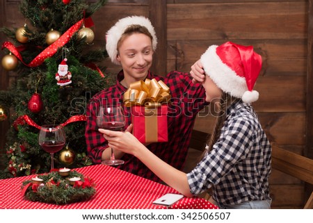 Man closing his girl-friend's eyes while she is holding a glass of red wine. Happy man going to present her New Year and Christman present.