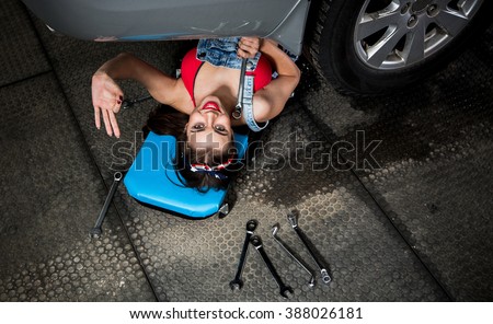 Young woman solving car problems while lying under car and fixing it in garage. Beautiful lady showing okay sign.