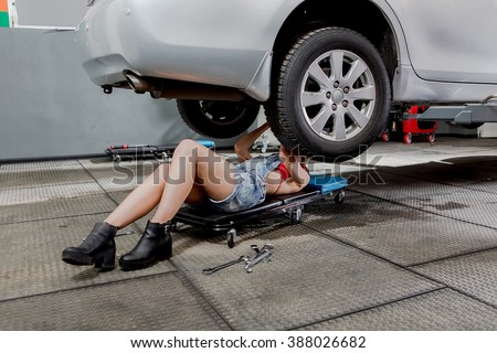 Mechanic woman underneath car checking the car conditions. Pretty lady in jeans shorts lying under car and repairing it.