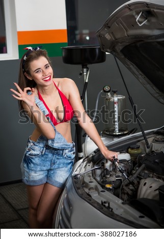 Photo of young female car repair worker. Glamour sexy brunette wearing jean shorts. Girl looking at camera, standing near car cowl and making okay gesture.