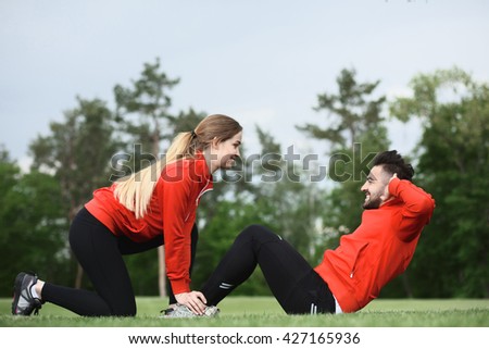 Sport lady holding man\'s legs who is doing abdominal crunches on fresh air in green park or forest. Fitness man preparing for competitions.