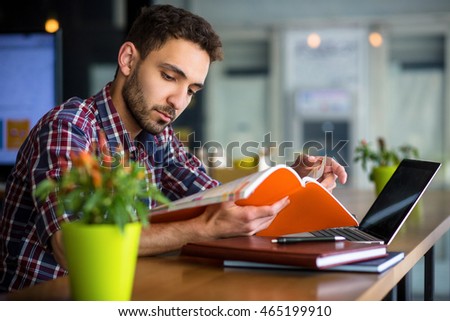 Portrait of handsome student reading book or document while studying in restaurant or cafe. Man using laptop computer for preparation to exams or classes.