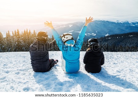 Three people in ski suits are sitting on the snow at the top of the mountain and looking into the distance. Family of skiers resting on the top of the mountain. back view