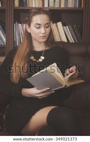 beautiful young girl in a black dress sitting on a brown leather couch with a book in the library.holding a book.read book