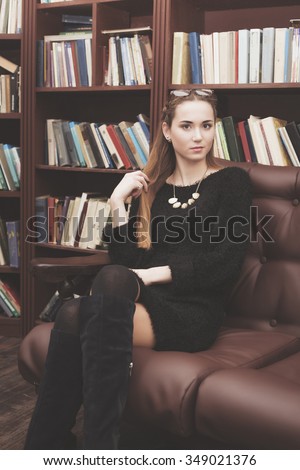 beautiful young girl in a black dress sitting on a brown leather couch with a book in the library
