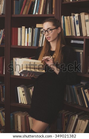 beautiful young girl in a black dress stands at the bookcase in the library.holding a book.read book