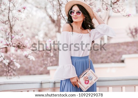 Outdoor portrait of young beautiful woman wearing stylish round sunglasses, straw hat, cold shoulder blouse, pleated skirt, with small white bag. Spring fashion concept. Copy, empty space for text
