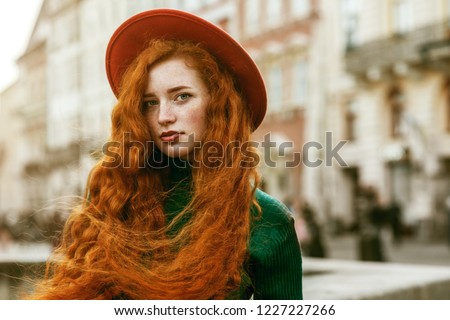 Close up portrait of young beautiful fashionable redhead woman with freckles, very long curly hair, wearing green turtleneck, orange hat, posing in street of european city. Copy, emty space for text