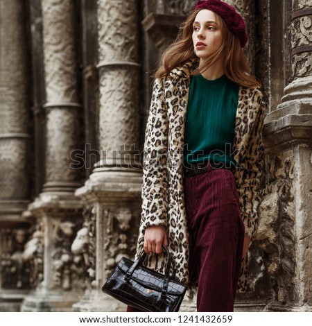 Outdoor fashion portrait of woman wearing trendy animal, leopard print faux fur coat, beret, sweater, corduroy trousers, holding  reptile skin textured bag, posing in street of city. Copy, empty space