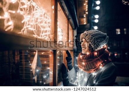 Outdoor night photo of young beautiful happy smiling girl looking through shop window with festive decoration, posing in Christmas fair, in street of european city, wearing knitted beanie hat, scarf